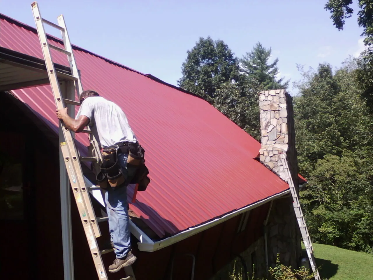 Roofer on a ladder beside a red metal roof and stone chimney — Soffit & Fascia Repair by Roofing Brothers in East Petersburg